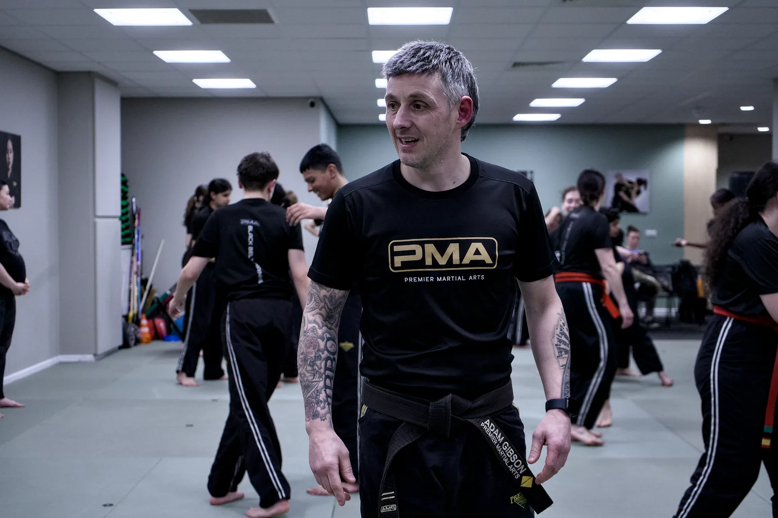 An instructor smiling on the mat with a class training behind