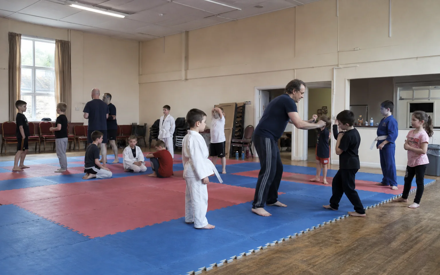 A mixed kids martial arts class on red and blue foam mats in a community hall